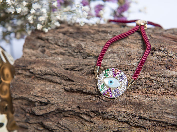 PULSO CORDON SEDA COLOR TINTO, AJUSTABLE CON TERMINALES Y DIJE CIRCULAR CON ZIRCONIAS Y OJO EN MADRE PERLA EN CHAPA DE ORO 14K